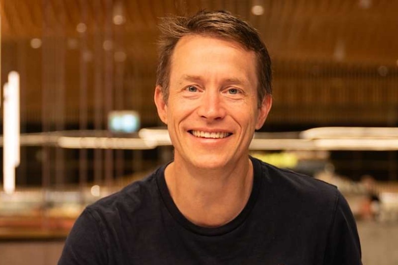 Smiling James wears black tee with sendler tag, background is blurred.