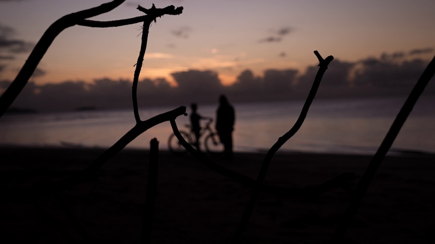 The silhouette of a boy on a bike and a woman on the beach during sunset. 