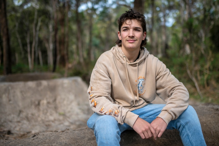 A teenager in a beige hoody and jeans sits on top of a steep bike jump he built from dirt in the bush.