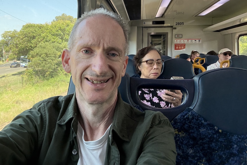 Mr Thomas wearing a green jacket with a white shirt smiles while on a train with people in seats behind him.