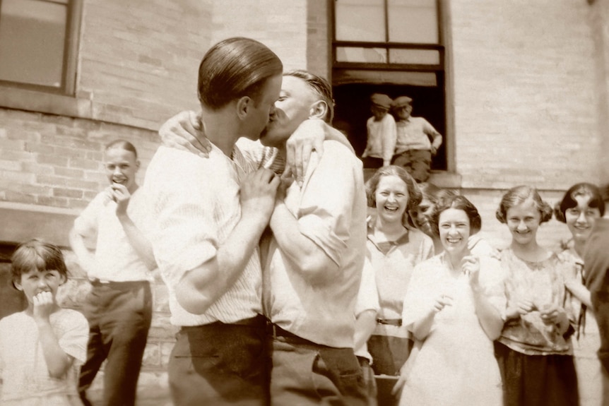 A black and white photo of two men kissing as people cheer.
