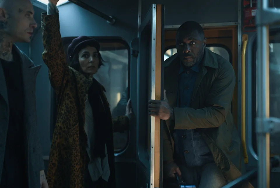 Idris Elba in Hijack. He is on a subway train grasping a pole next to two other passengers.