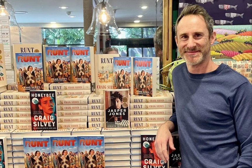 Craig smiles next to a huge stack of books including Runt, Honeybee and Jasper Jones