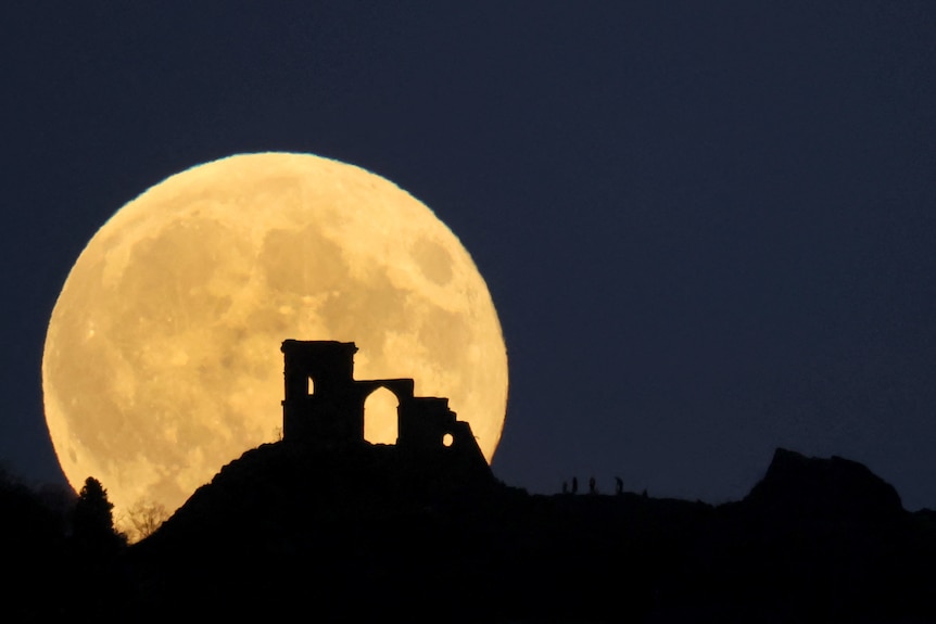 A full moon rises above a hill with the ruins of a castle on it