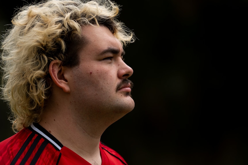 A man with blonde hair against a dark background looking into the distance.