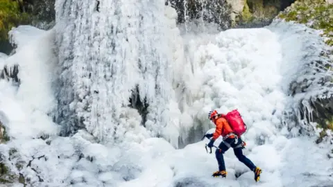 PA Media A man wearing a bright red jacket and yellow and black hiking boots climbs across a frozen waterfall