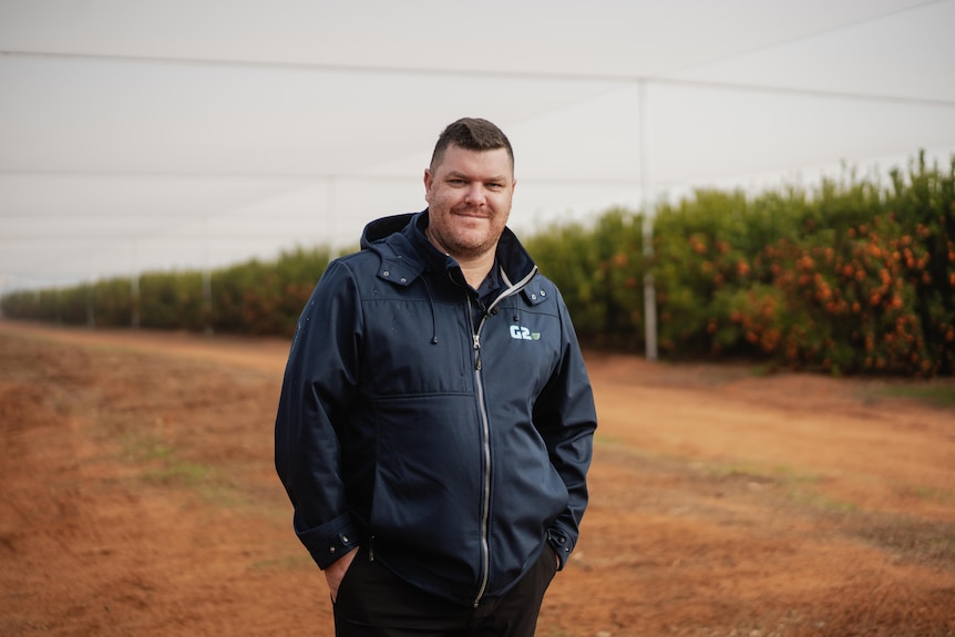 a man smiles at the camera in front of orange trees.