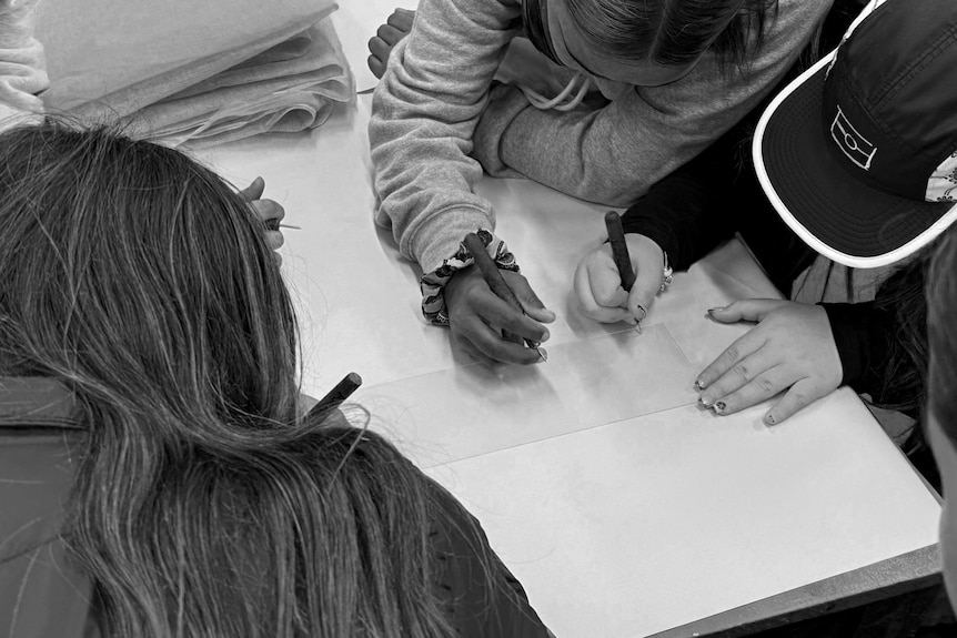 A black and white photo looking down on three young Indigenous people etching an artwork.