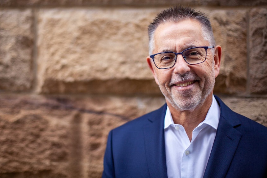 Norman Swan is wearing a blue suit and standing in front of a sandstone brick wall.