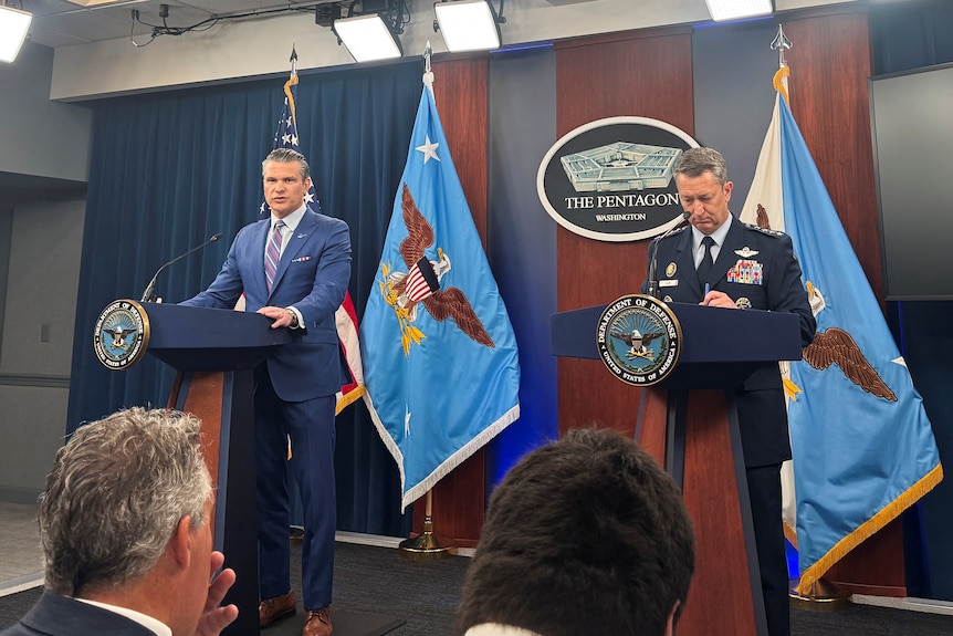 Two men in suits stand behind lecterns with flags in the background.