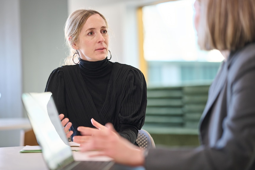 Nicola Henry wearing a black sweater sitting down at a table speaking with someone