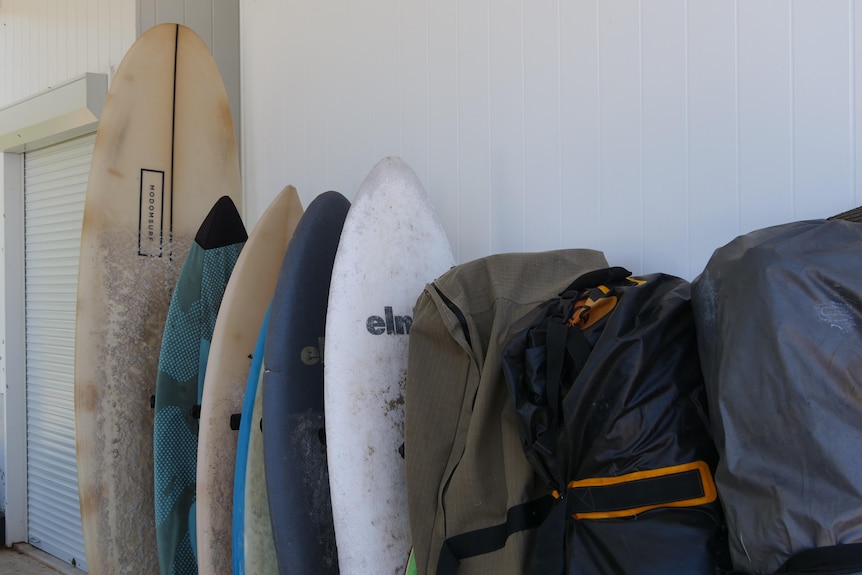 Rack of surfboards stacked vertically against the wall of a house, with some bagged.