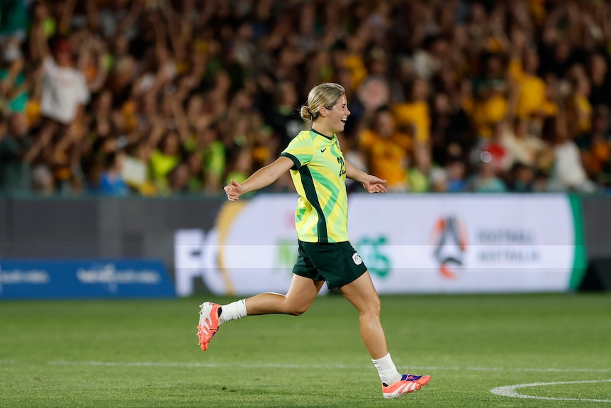 A soccer player in green and gold runs arm outstretched and smiling