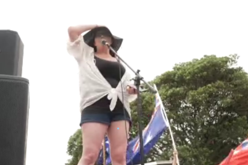 A woman wearing a hat standing next to speakers and australian flags speaking into a microphone