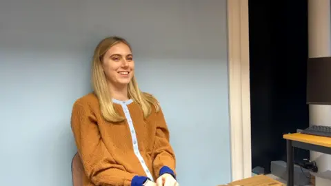 Sadie Rees a young blonde haired girl sat in a chair against a wall smiling.