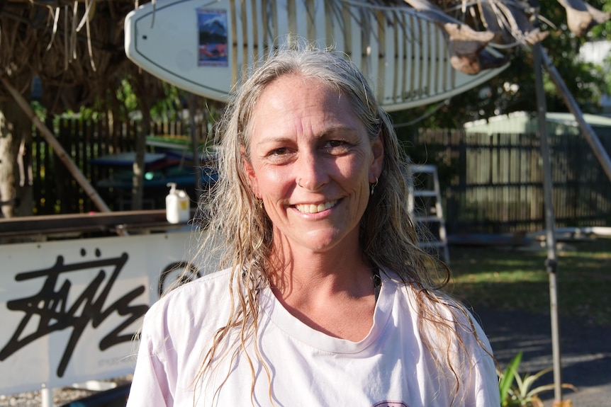 Woman with long light coloured hair, wearing a t-shirt and smiling at the camera with a surfboard in the background.