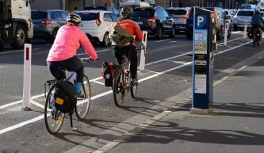 Three people ride on a painted bike lane with white bollards in the road to their left, with a blue parking meter on the footpath to their right and traffic in the road alongside the lane.