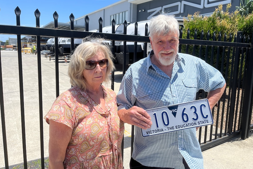 upset couple holding number plate outside gates with caravans in background