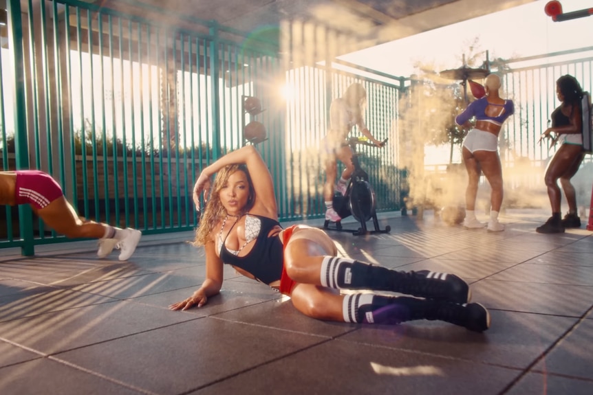 Tinashe lies on a gym floor with other women working out around her
