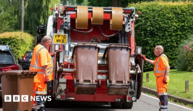 Two men in orange hi-viz overalls stand either side of the back of a waste collection vehicle. The truck is in the process of emptying two brown bins. Behind the van there is a patch of grass and several large green bushes, as well as a couple of parked cars on the road.
