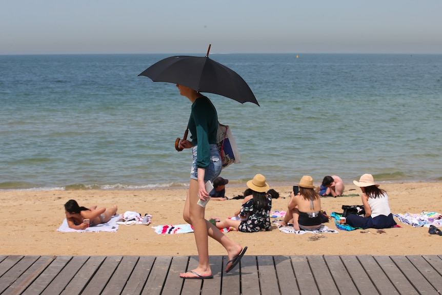 A woma  walks along a sandy beach in bathers holding an umbrella