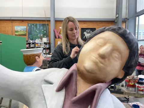 A woman paints the dark hair of the sculpture Tom who is dancing. He does not have any features yet. The face is still to be painted