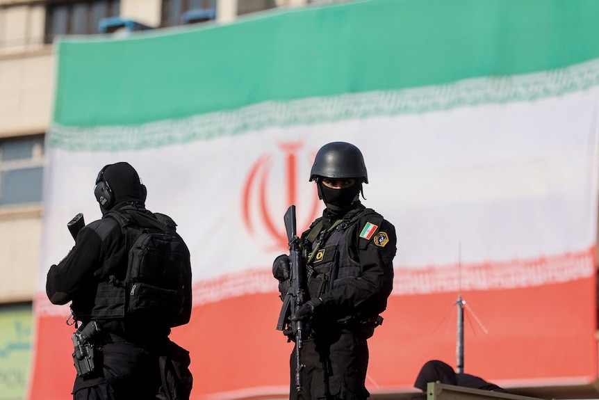 Two heavily armed policemen stand in front of an Iranian flag