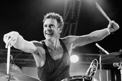 A black and white shot of a man's arms in the air with drum sticks, sitting behind a drum kit, smiling and looking to the side.