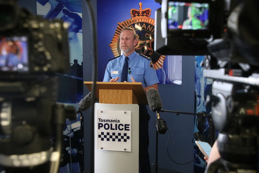 A man in a navy blue police uniform stands at a lectern surrounded by microphones, while the media films.