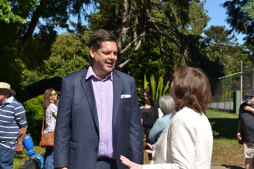 Tall man, with short dark hair, wearing a suit speaks to a woman with her back to the camera. 