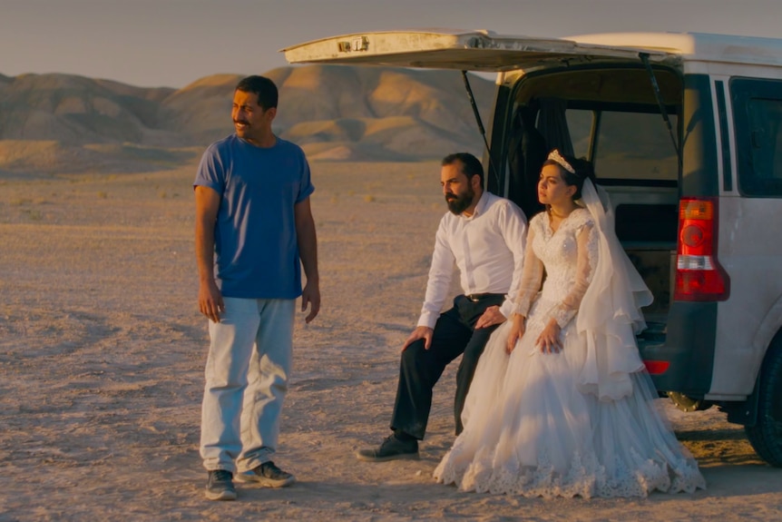 A bride and groom sit in the back of a van next to another man in the desert