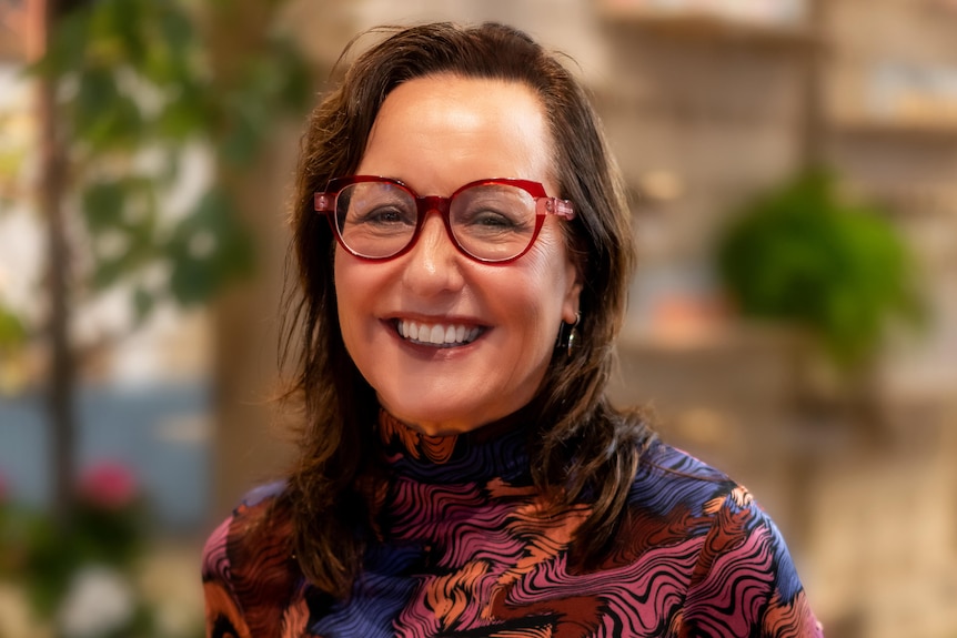 A woman wearing a pattern dress and glasses, smiling at the camera.