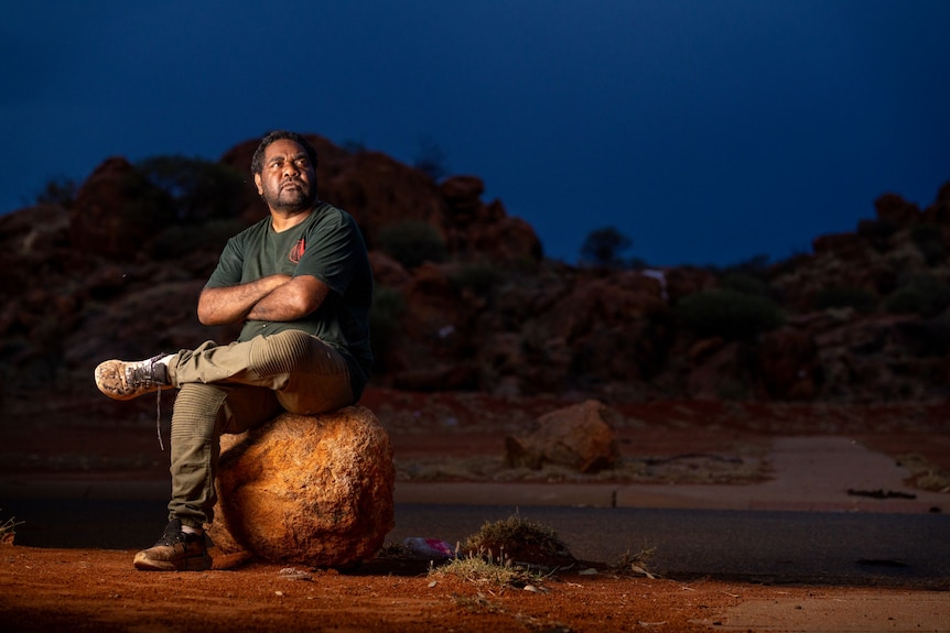 Artist Shane Dodd sits on a rock with his legs closed. It is night time. 