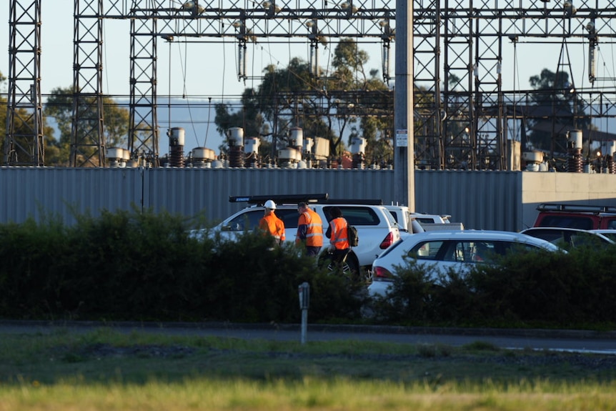 Workers wearing high-visibility clothing walk near parked cars in front of industrial power infrastructure.