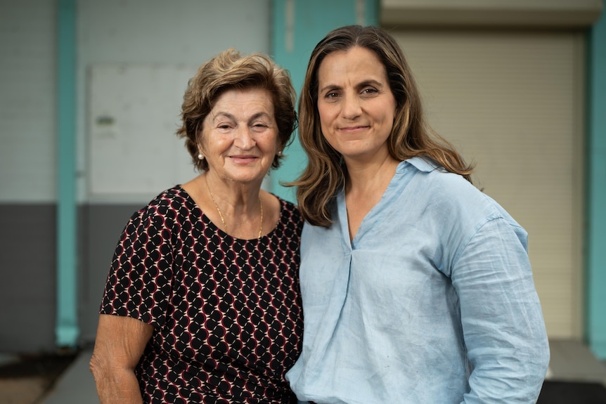 Adult mother and daughter standing together on a street side. Mum has short brown hair, pattern top, daughter in blue blouse