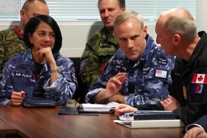 A woman in a military uniform sitting at a table and looking on, surrounded by multiple men in uniform