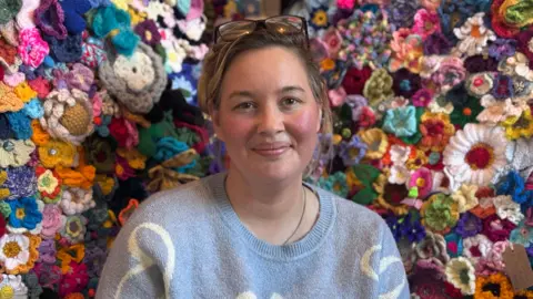 Tasha Lloyd sitting in front of a wall of thousands crochet flowers, each one different and brightly coloured. Tasha has brunette hair has glasses on her head. She is wearing a blue jumper and has a slight smile. 