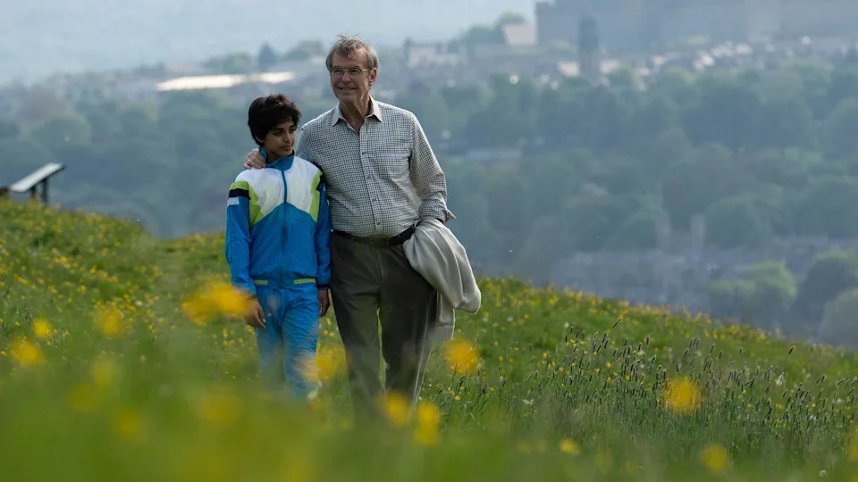 An elderly man in a smart shirt strolls through a flowery meadow on a Summer's day. His arm is affectionately placed around the shoulders of a young boy dressed in a blue tracksuit