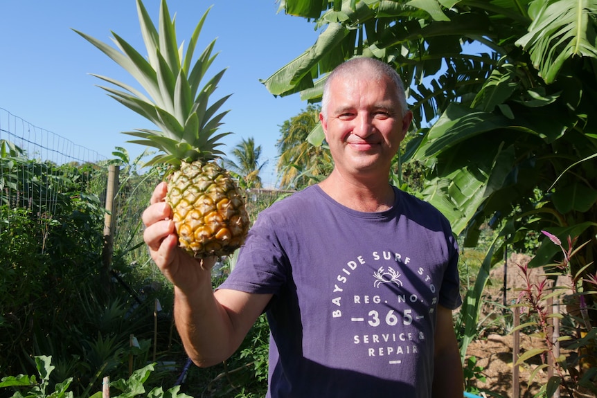 A man smiles and holds a pineapple up with a garden behind him.