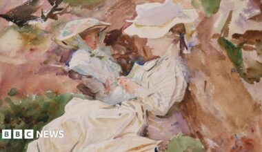 A man in a colourful red shirt with black glasses and long grey hair smiling at camera infront of artwork