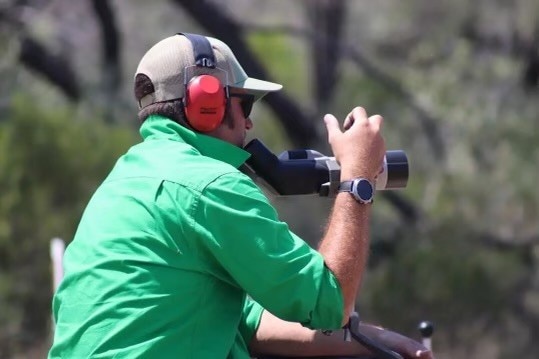 A man in a green shirt with ear muffs on looks though a scope.