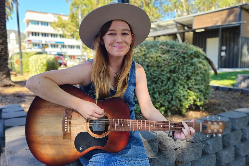 A long haired woman in a blue dress and broad brimmed hat plays guitar in a park