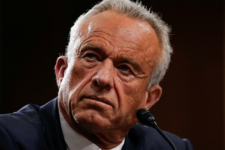 A clean-shaven, tanned man with short, white hair and a serious expression.