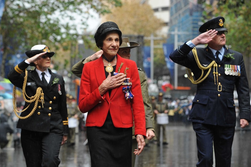 A woman in a brightly-coloured blazer puts her hand to her heart as she marches alongside uniformed personnel down a city street