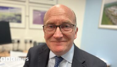 Nick Hulme standing in the elderly ward at Colchester Hospital shortly before he retired at the end of December 2025. He is wearing a lanyard around his neck that describes his position with a name badge saying "Nick". He has a pale blue and white pinstripe shirt and a blue tie. He is wearing thick plastic black glasses and has a short beard. In the background you can see beds within the ward and a dirty bed sheet trolley. The walls are yellow and he is standing to the side of some red cabinets and monitors.
