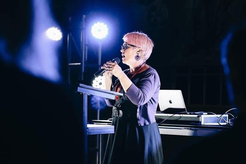 A woman with short pink hair stands on a stage, speaking into a microphone, bright lights behind her