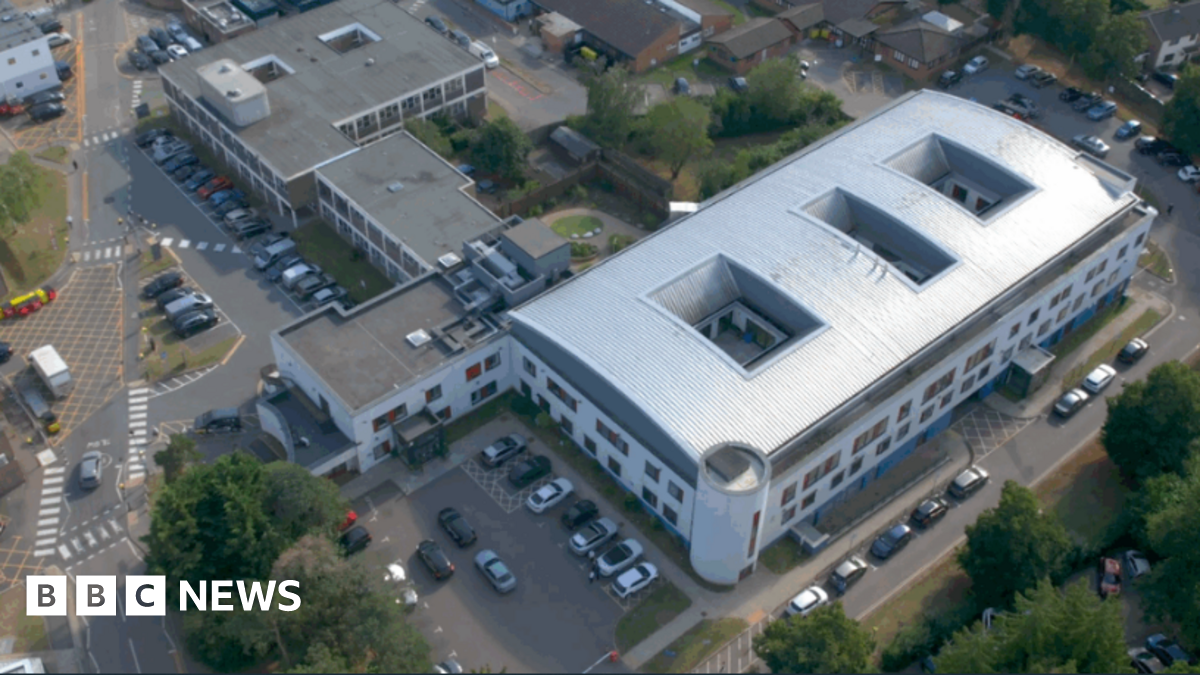 An aerial view of the large Derwent Centre complex in Harlow. It features a modern, white, curved-roof structure at the centre. Surrounding it are several older, rectangular buildings arranged along internal roads and parking areas. Cars are parked in multiple spaces and the site is bordered by trees.