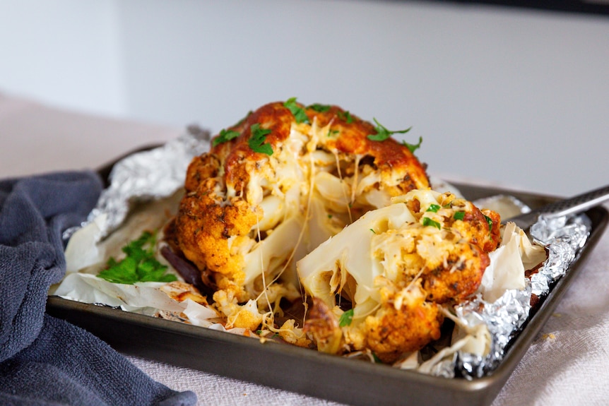 A whole roasted cauliflower is cut open after baking, revealing melted cheese. It's topped with chopped parsley.