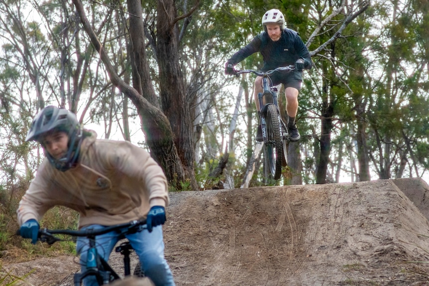 Two mountain bikers catch air over steep dirt bike jumps in the bush.