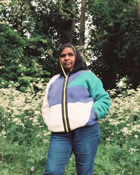 Portrait of Britain/Anupama Gamanagari Anupama Gamanagari's mother stands in the Staffordshire countryside. She is wearing a purple, white and green fleece with the black hood up. She is also wearing jeans and stands amid greenery in front of trees. 